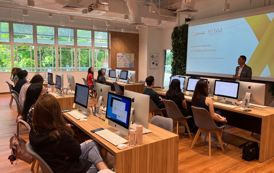 A presenter leads a Bot Building Workshop in a modern computer lab with rows of iMac desktops and seated attendees.